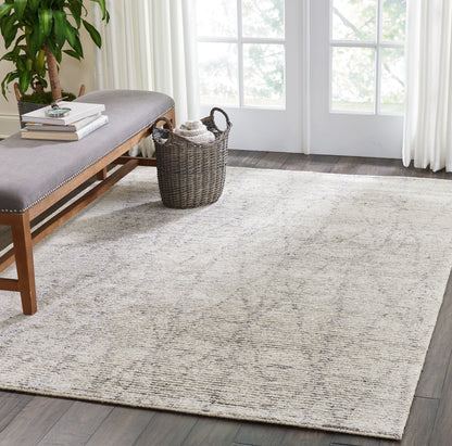 Beige textured area rug on dark wood floor with gray upholstered bench, wicker basket, potted plant, and large windows.