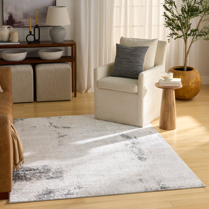Beige fabric armchair with dark gray pillow, wooden stump side table & large plant, neutral abstract rug, warm wood floors.
