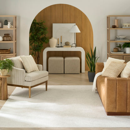 Neutral living room with tan leather sofa, cream chairs, white rug, wood arch backdrop, plants & shelves.