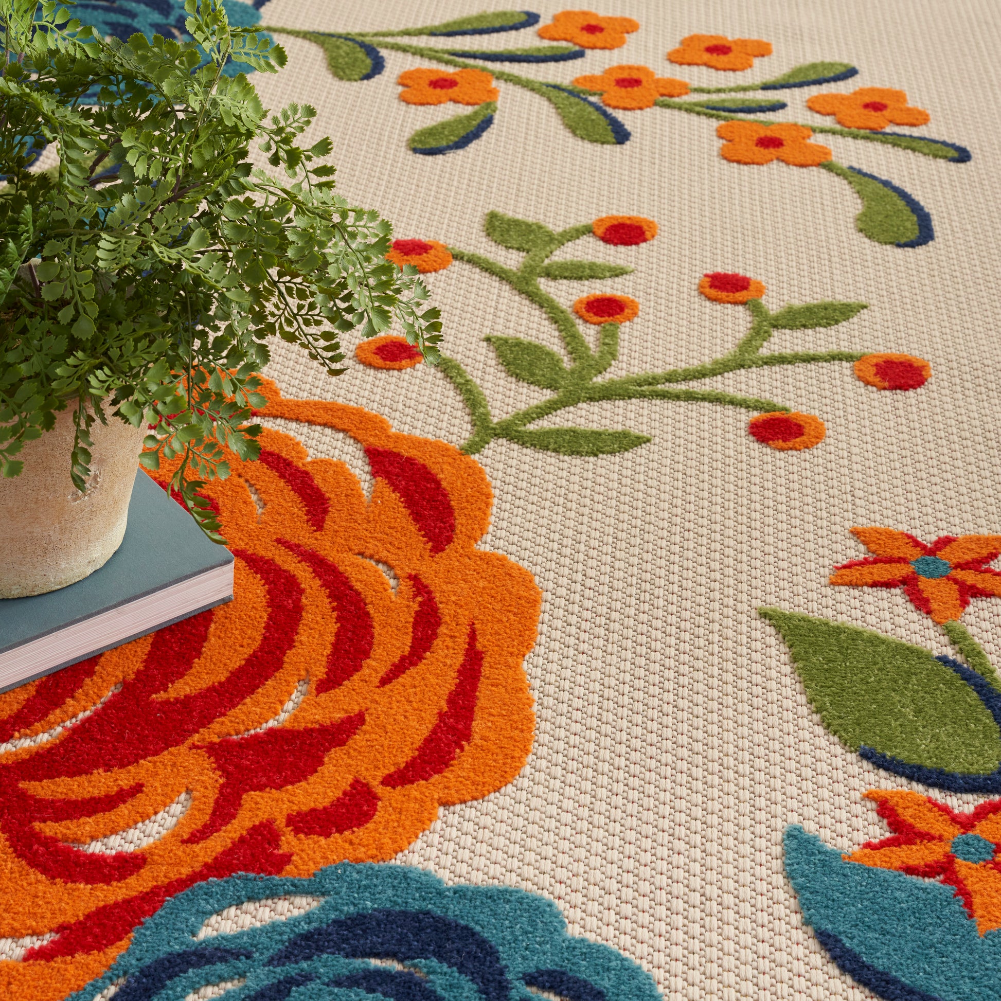 Beige woven area rug with vivid orange and blue floral pattern; potted fern rests on left edge.