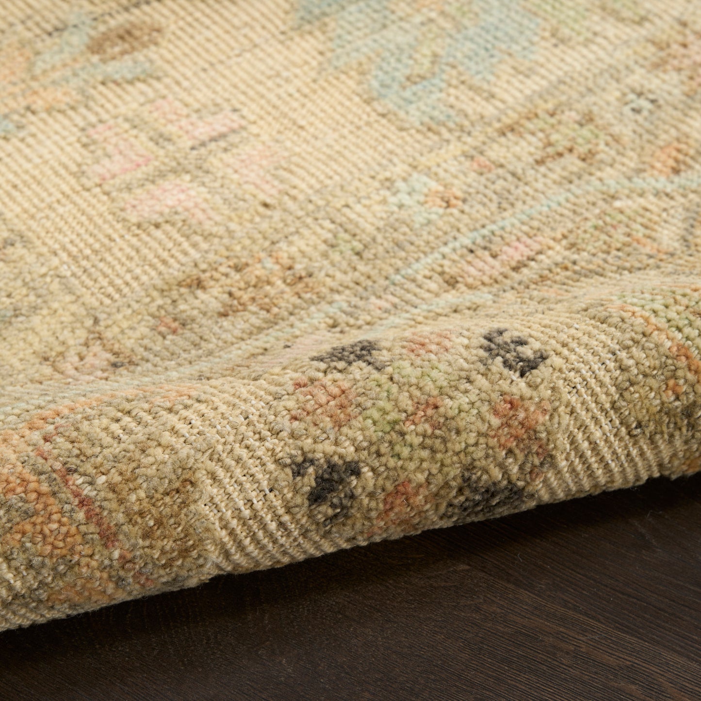 Beige woven rug with pastel blue, pink & green floral motif; folded edge on dark wood floor.