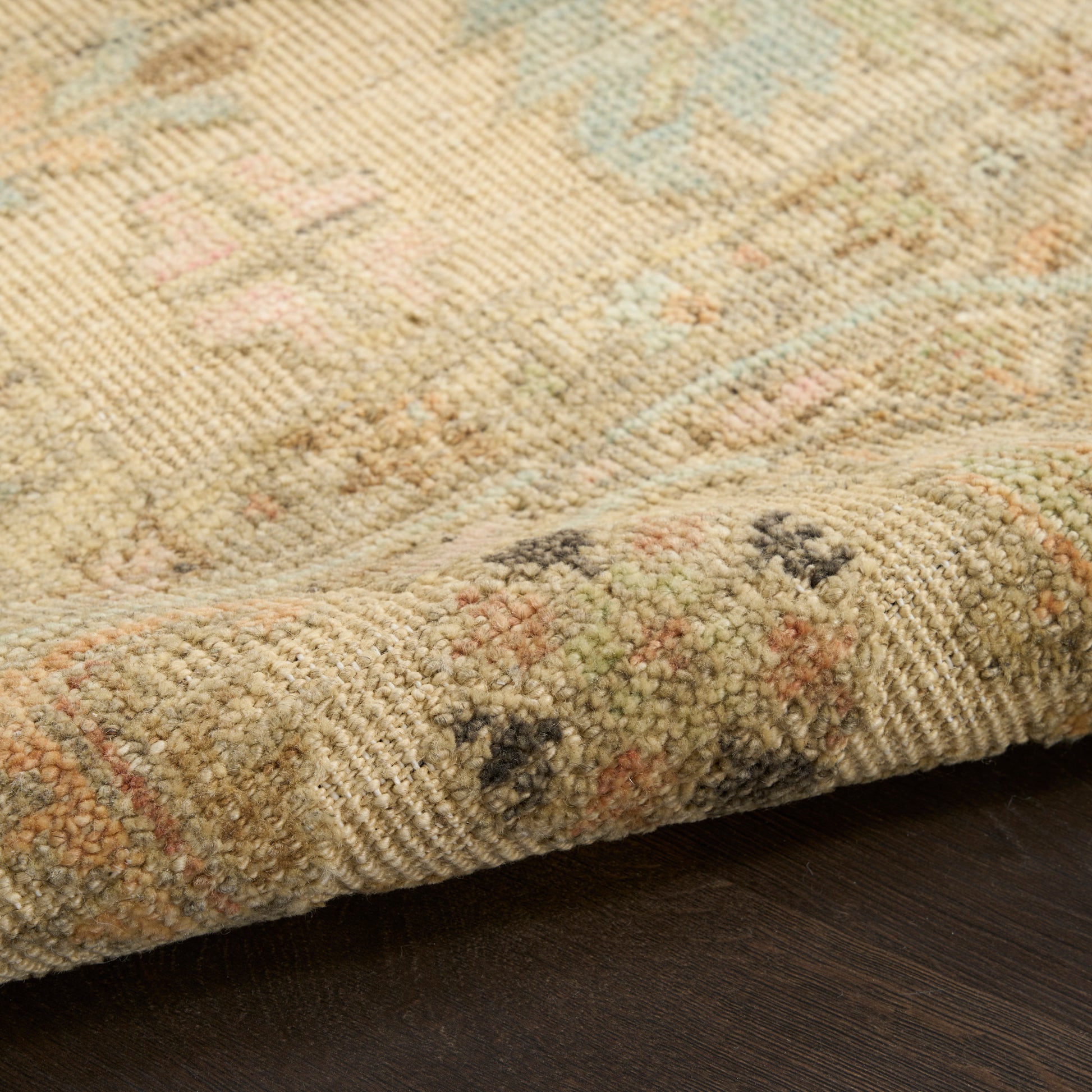 Beige woven rug with pastel blue, pink & green floral motif; folded edge on dark wood floor.