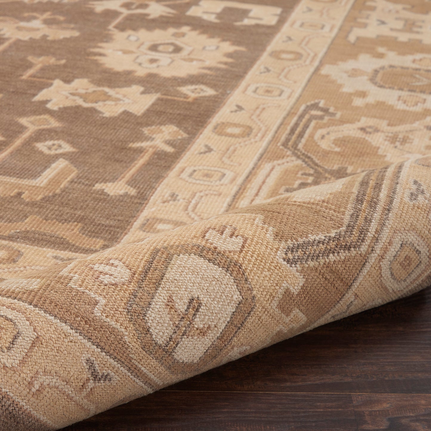 Beige-brown traditional oriental rug with geometric motifs; folded corner on dark wood floor.
