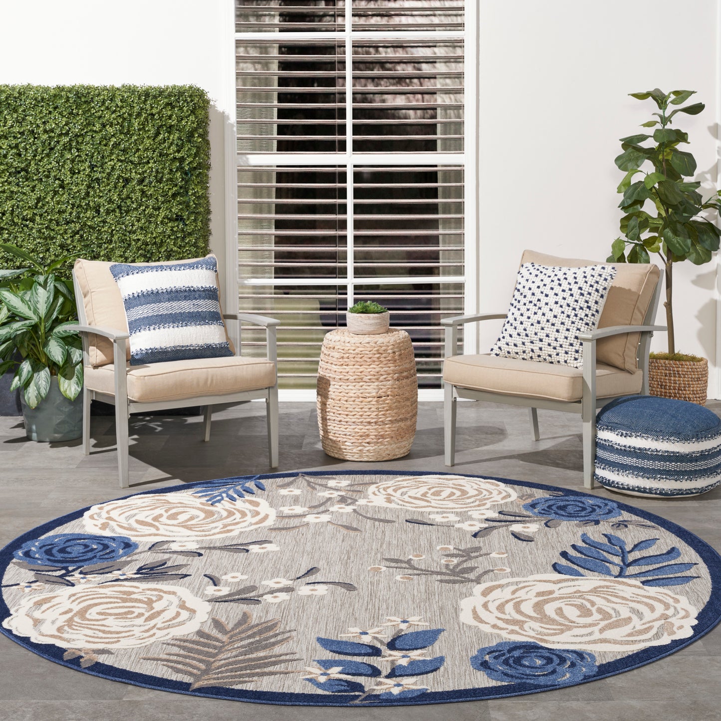 Round beige-blue floral outdoor rug with two beige cushioned chairs, blue pillows, wicker side table & potted plants.