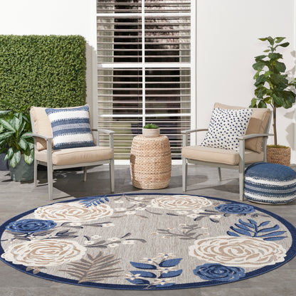Round beige-blue floral outdoor rug with two beige cushioned chairs, blue pillows, wicker side table & potted plants.