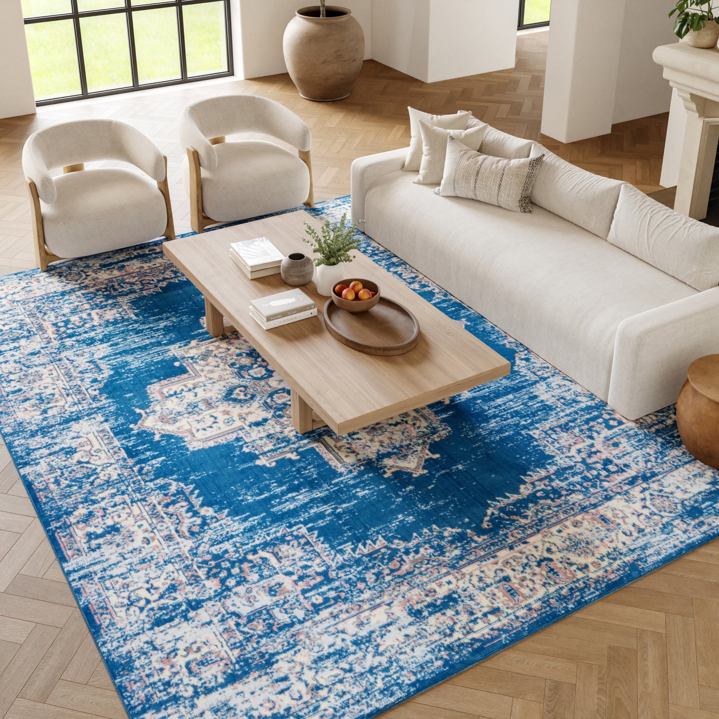 Living room with blue distressed rug, beige sofa, two cream curved armchairs, and light wood coffee table.