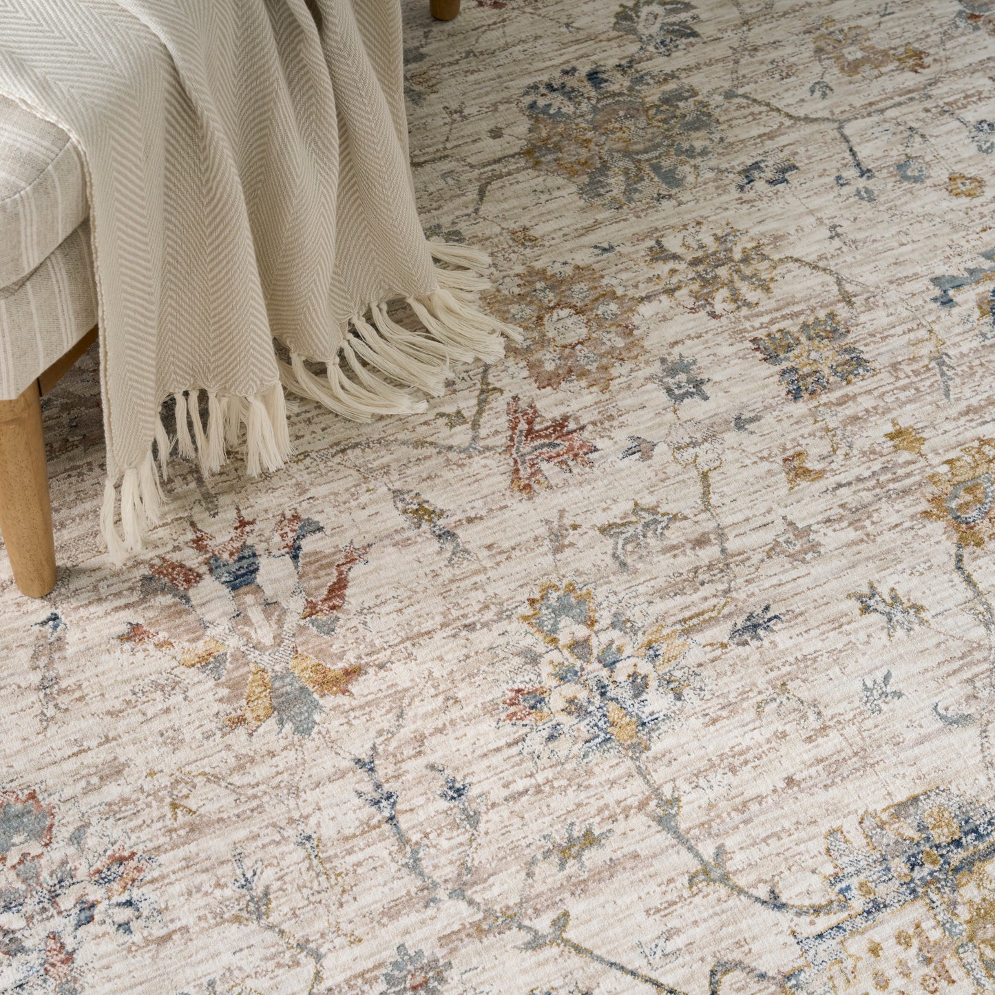 Cream fringed throw over a light sofa, atop a beige distressed floral rug with pastel blue, yellow and orange accents.