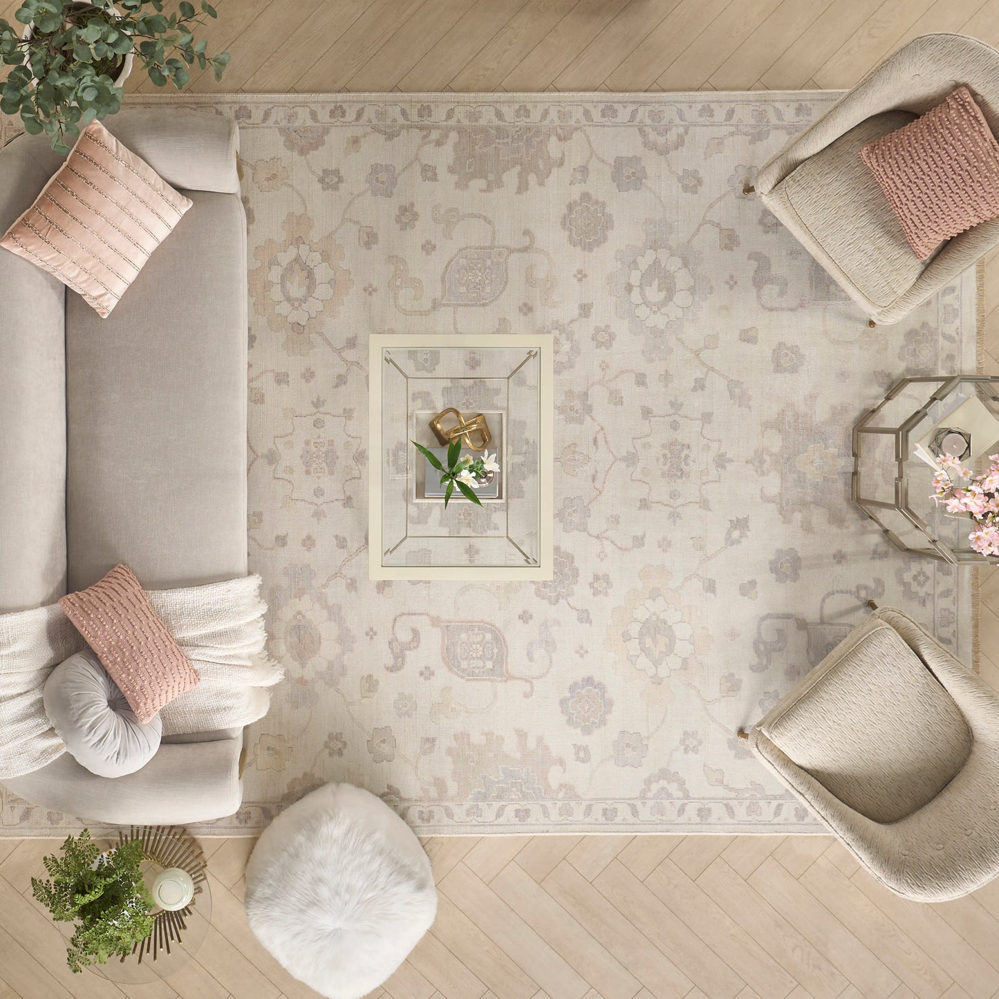 Cream sofa with blush cushions, white round pouf, light armchairs, glass coffee table, and beige floral rug.