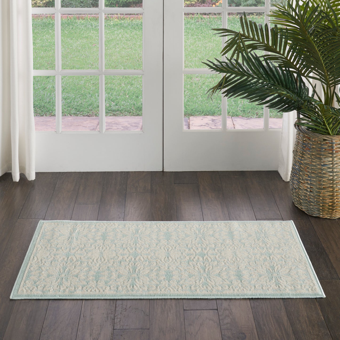 Rectangular cream rug with pale teal border and subtle pattern on dark wood floor near glass doors, with potted plant nearby.