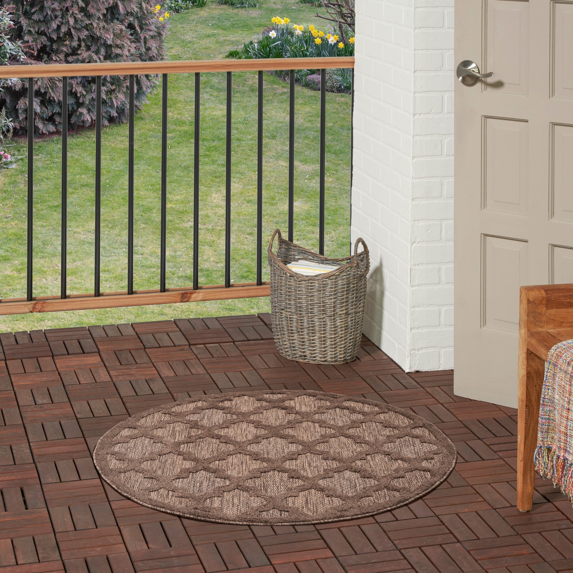 Round brown braided outdoor rug on a wooden deck beside a wicker basket and doorway.