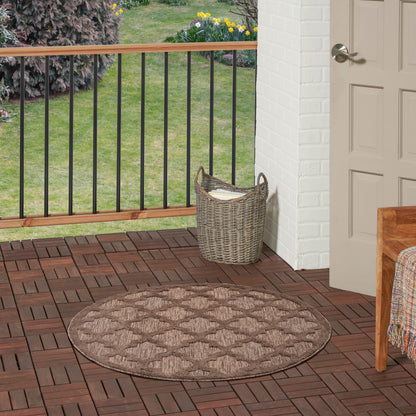 Round brown braided outdoor rug on a wooden deck beside a wicker basket and doorway.