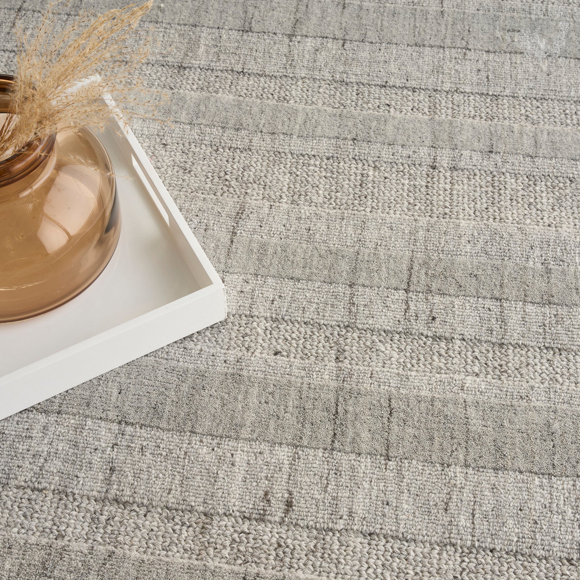 Brown rounded glass vase with dried pampas grass on a white tray atop a gray woven rug.