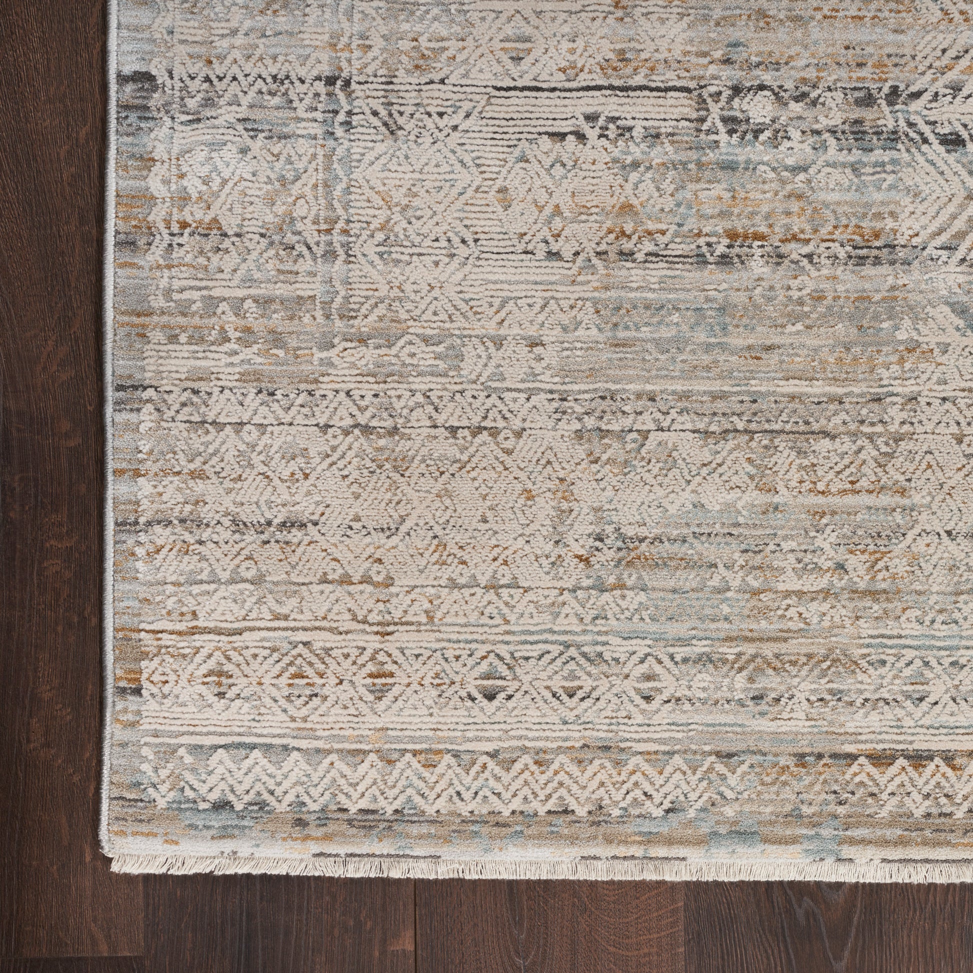 Distressed geometric area rug in ivory, taupe, gray and blue on dark wood floor; fringed edge.