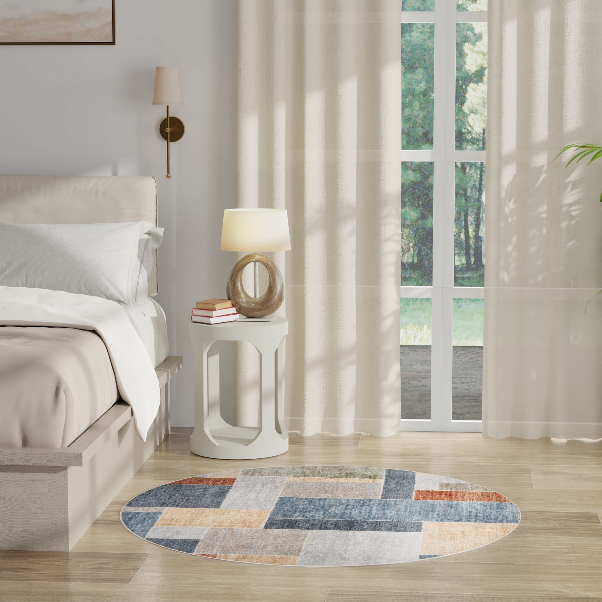 Cream bed with white linens, round white side table with lamp and books, patchwork round rug, sheer curtains, glass doors.