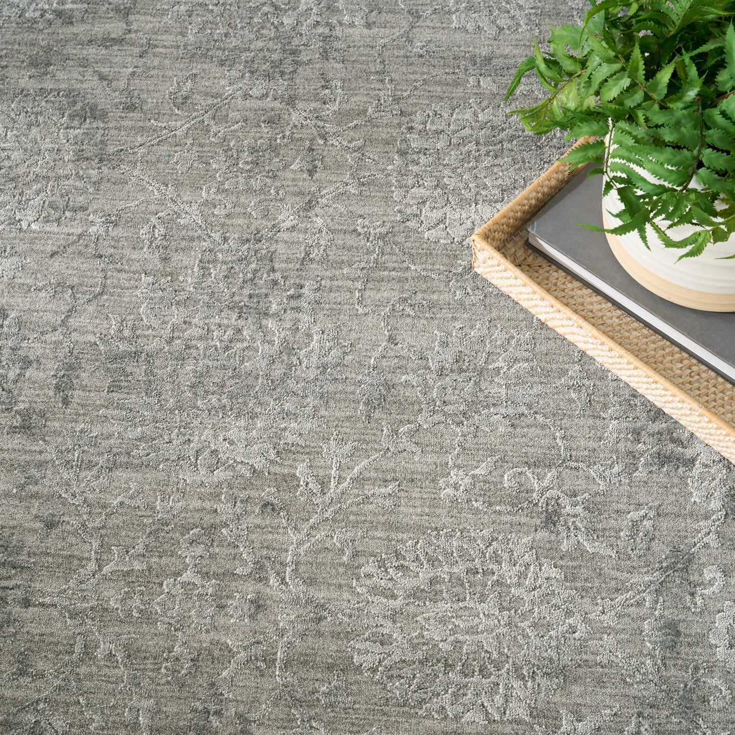 Gray patterned area rug with a damask design; wicker tray holding a potted fern & books in the corner.