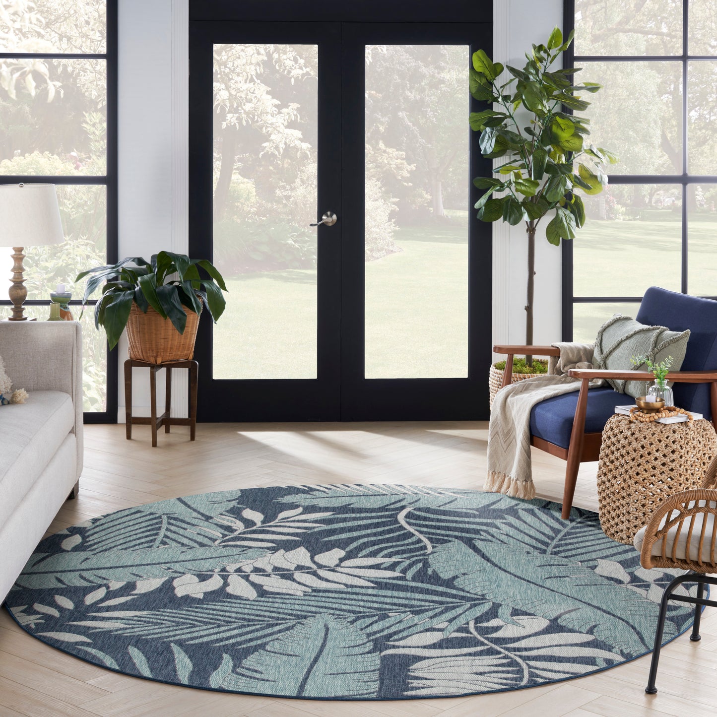 Round teal-blue tropical leaf outdoor rug in a sunny outdoor space with plants and woven furniture.