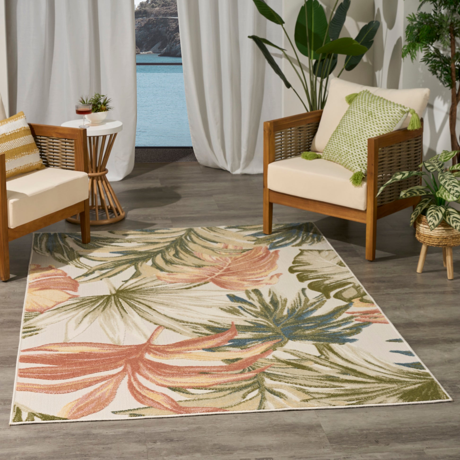 Palm leaf rug in pink, green and blue on a wooden floor, with beige cushioned chairs and tropical plants.