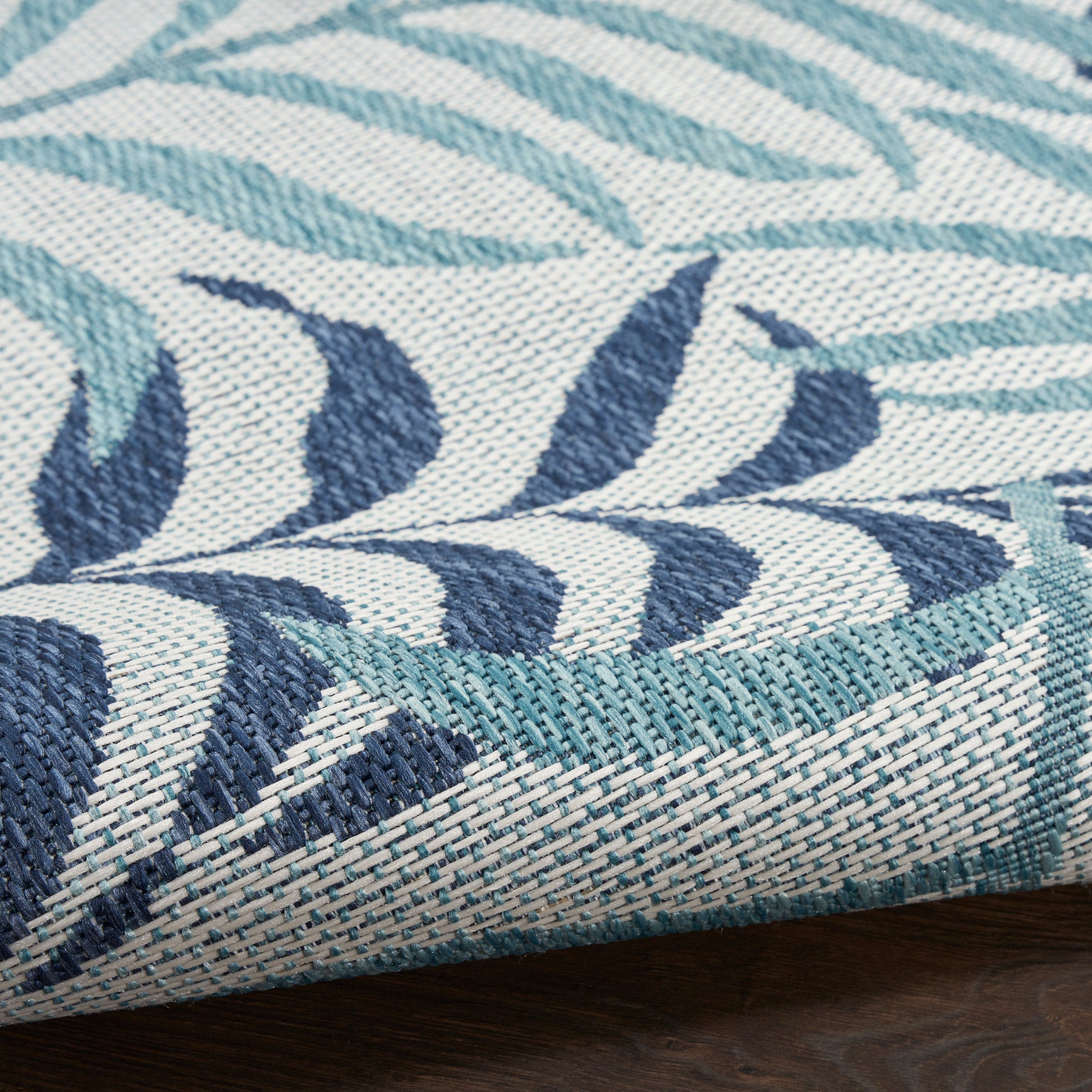 Woven rug with teal and navy leaf pattern on ivory background; textured weave with a folded corner.