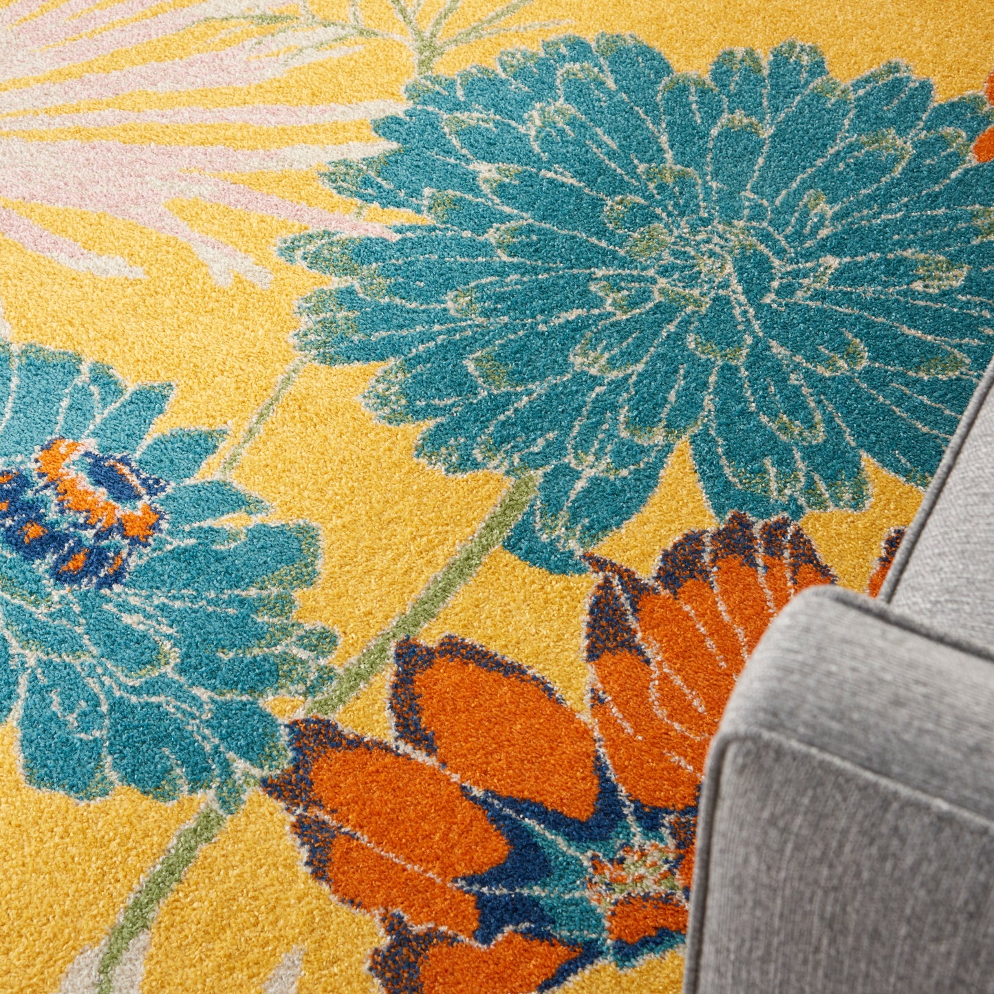Yellow floral rug with oversized teal-blue blossoms and orange accents beside a gray sofa.