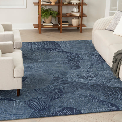 Blue fern-pattern area rug on light hardwood floor, flanked by beige sofas and a wooden shelf in a bright living room.
