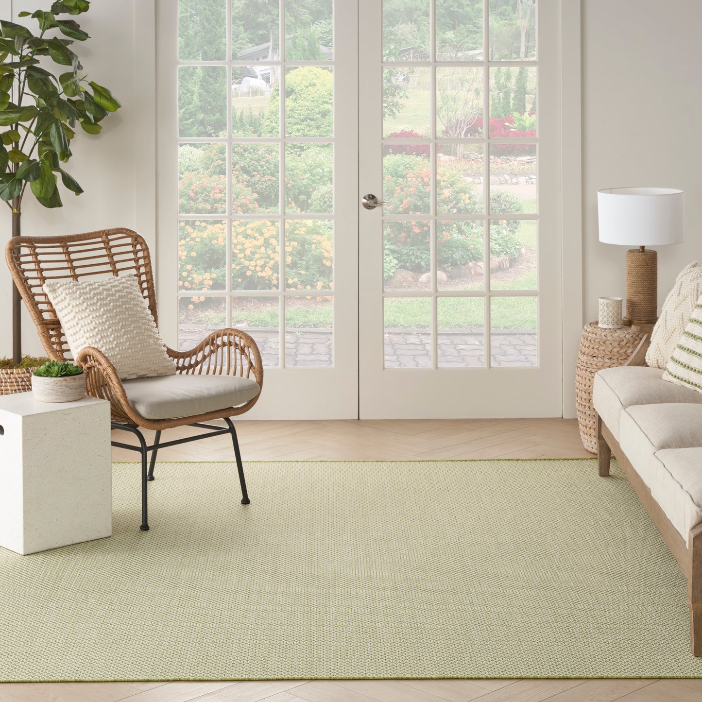 Coastal living room with a rattan chair, cream cushions, light green rug, beige sofa, and woven side table by French doors.