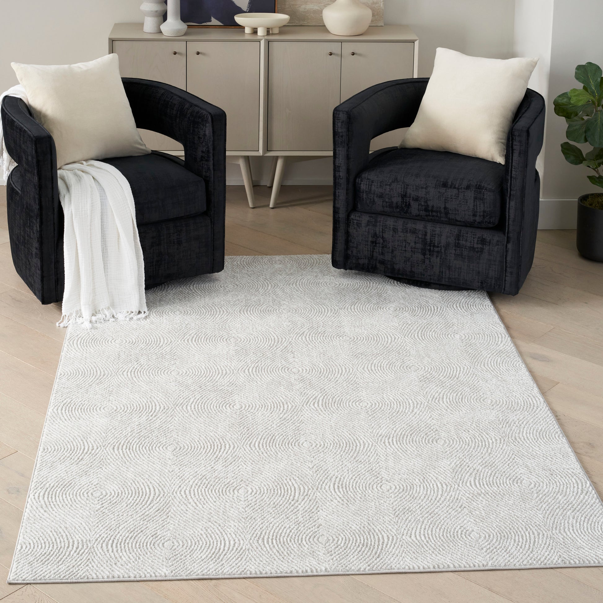 Pair of dark charcoal velvet armchairs with cream cushions and a white throw, on light patterned rug in modern living room.