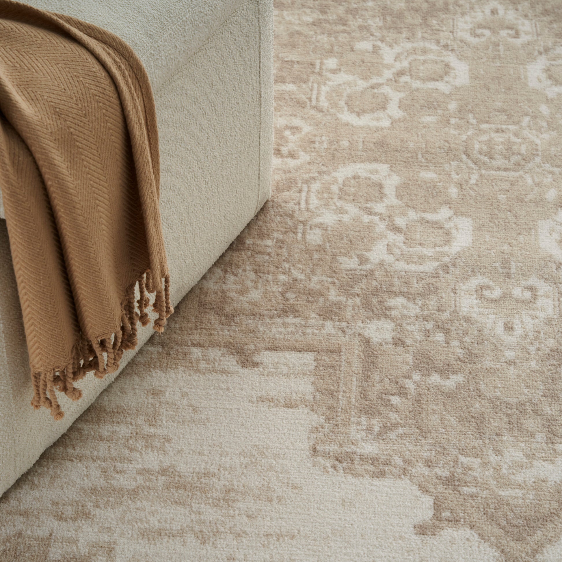 Beige sofa with tan fringe throw draped over edge, atop a light patterned neutral area rug.