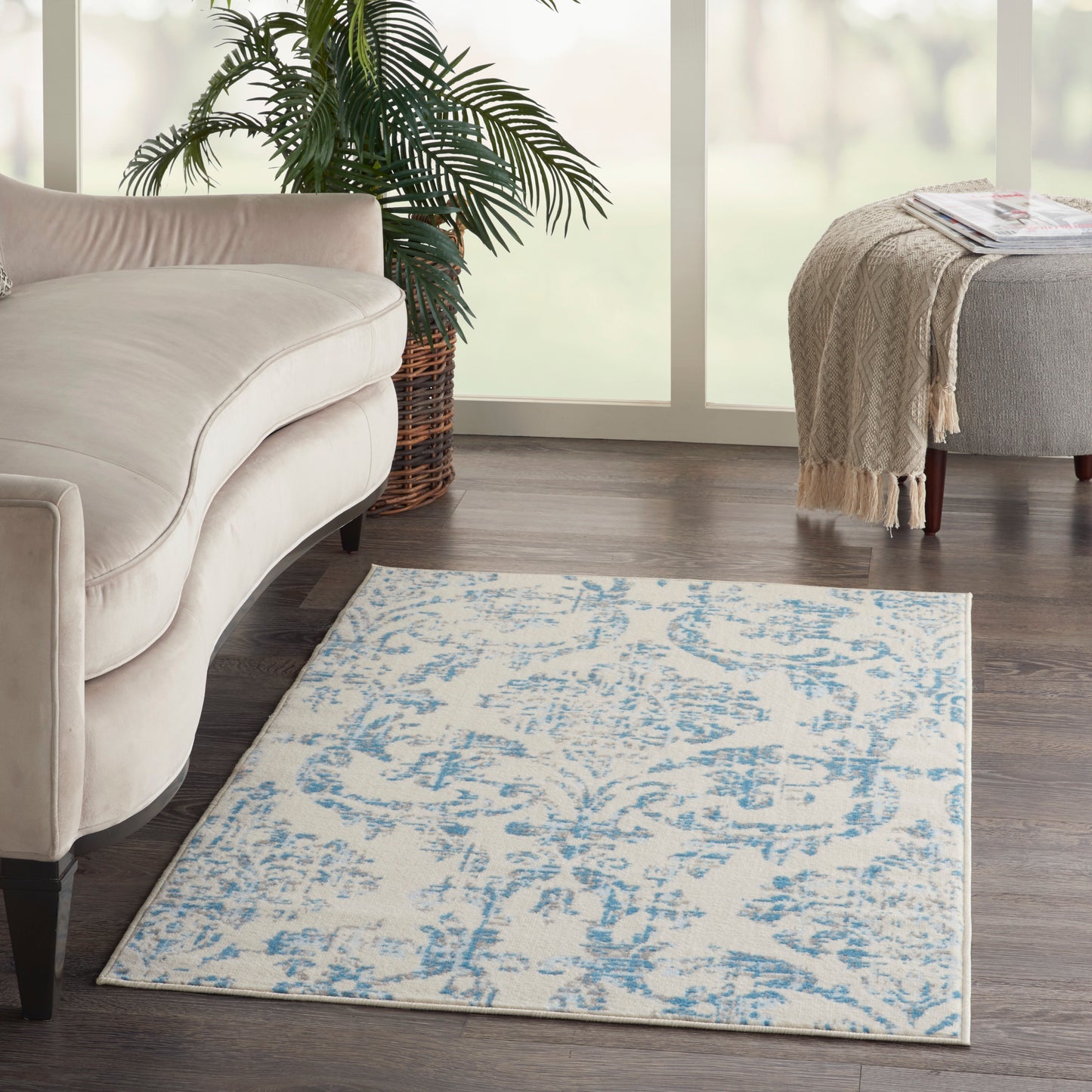 Cream curved sofa beside a blue patterned area rug, dark wood floor, green palm in wicker planter, beige ottoman with fringed throw.