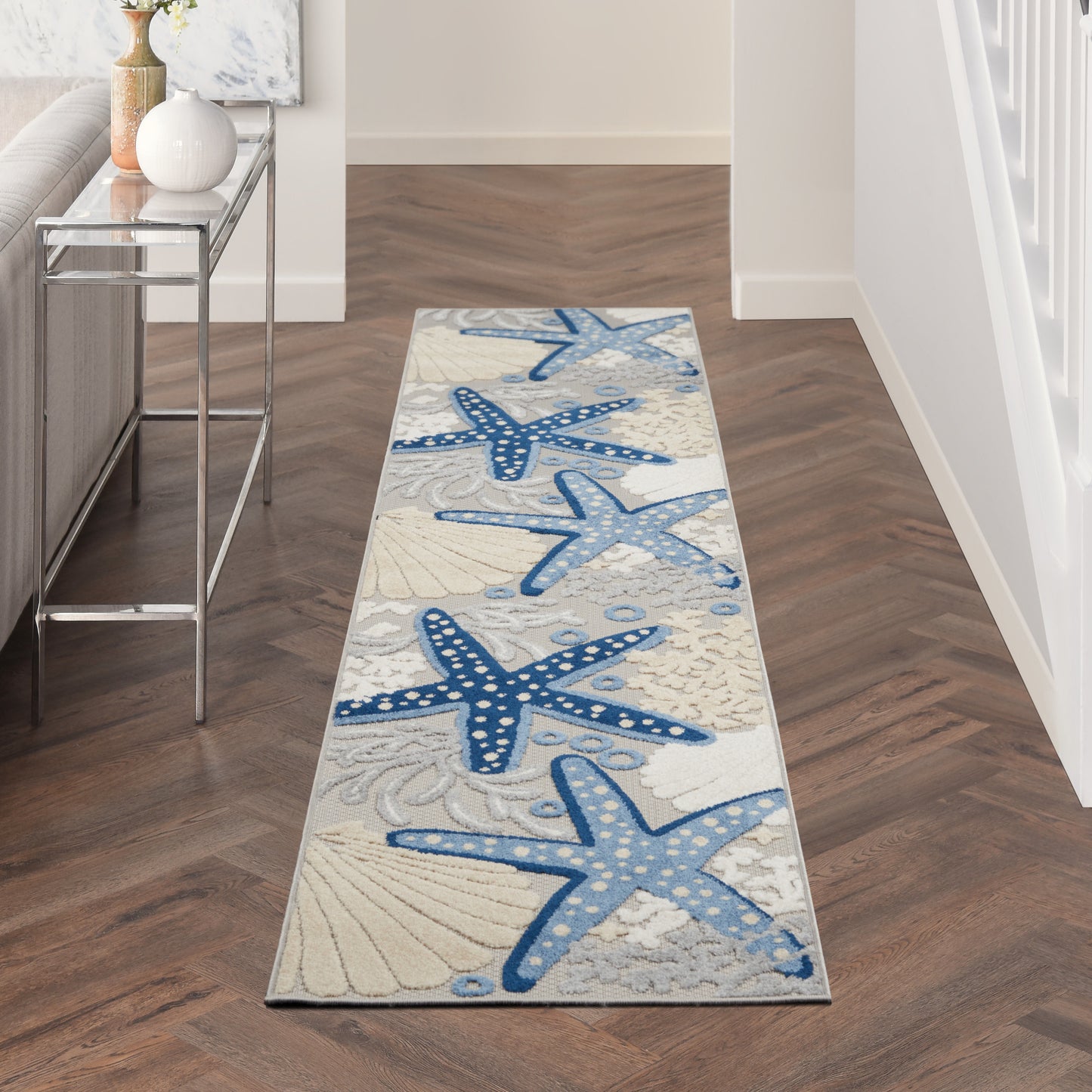 Long nautical runner rug with blue starfish motifs on a beige background in hallway beside a glass console table.