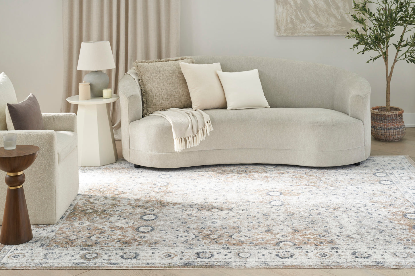 Light beige curved fabric sofa with cream and taupe cushions, a soft throw, in a neutral living room with a patterned rug.