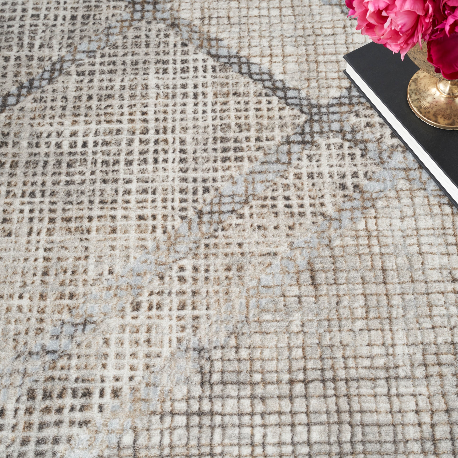 Beige patterned area rug with brown and gray grid design; pink peony bouquet in brass vase on a black notebook in corner.