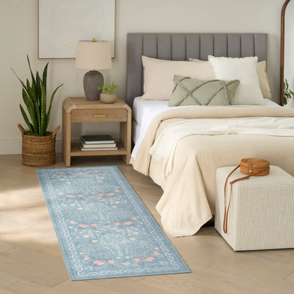 Neutral bedroom featuring a gray upholstered headboard, beige bedding, blue floral runner rug, wooden nightstand, snake plant, and an ottoman.