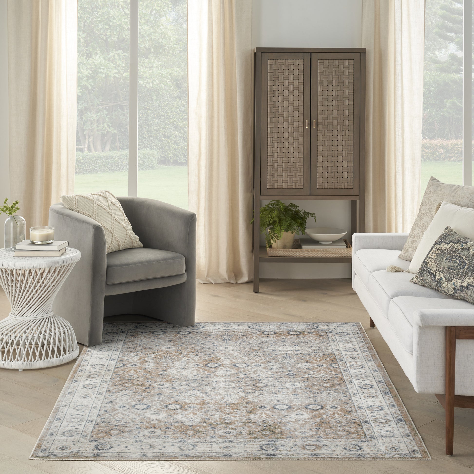 Bright living room with grey armchair, cream sofa, woven-paneled cabinet, potted plant, and a blue-gray patterned area rug.