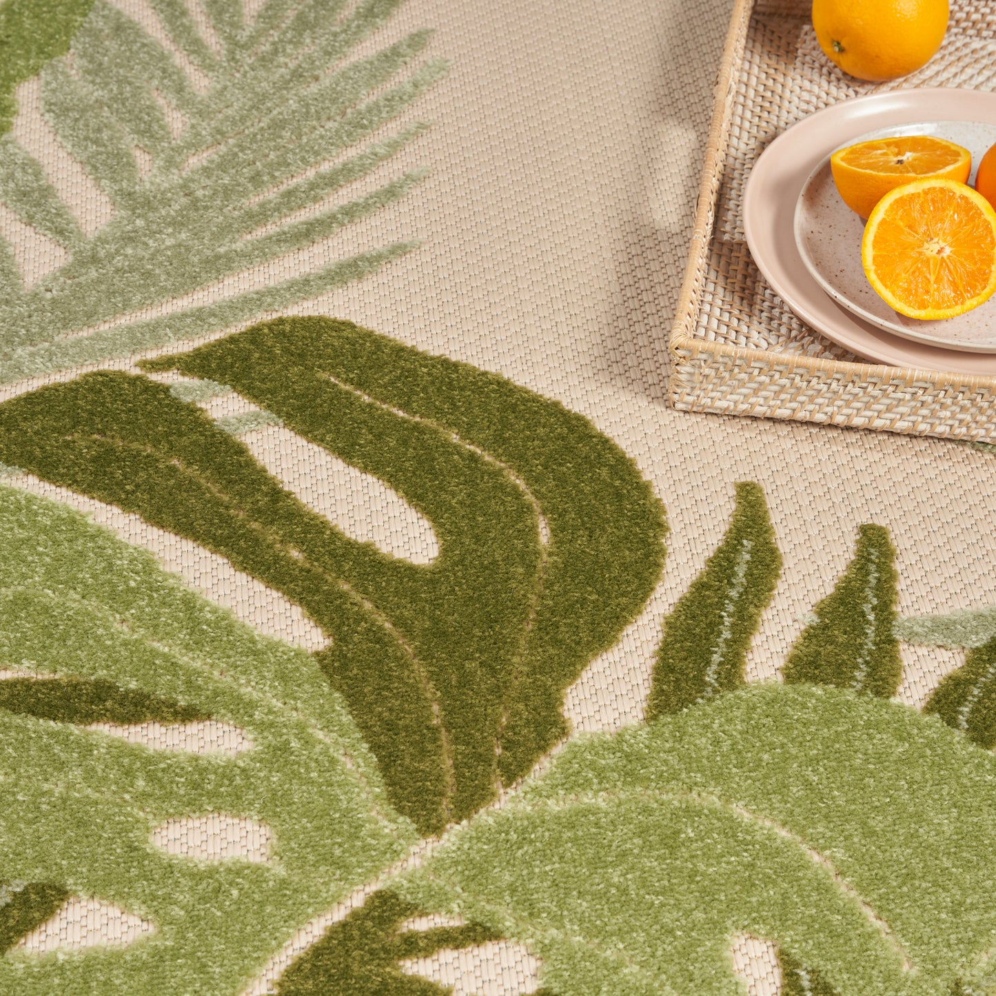 Beige woven outdoor rug with a large green monstera leaf pattern; wicker tray with orange halves on a plate.