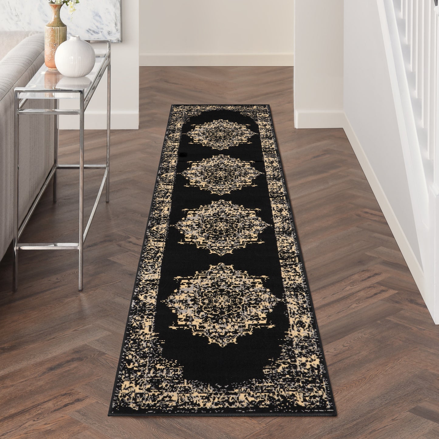 Long black hallway runner rug with beige medallions and border on a dark herringbone floor beside a silver metal console.