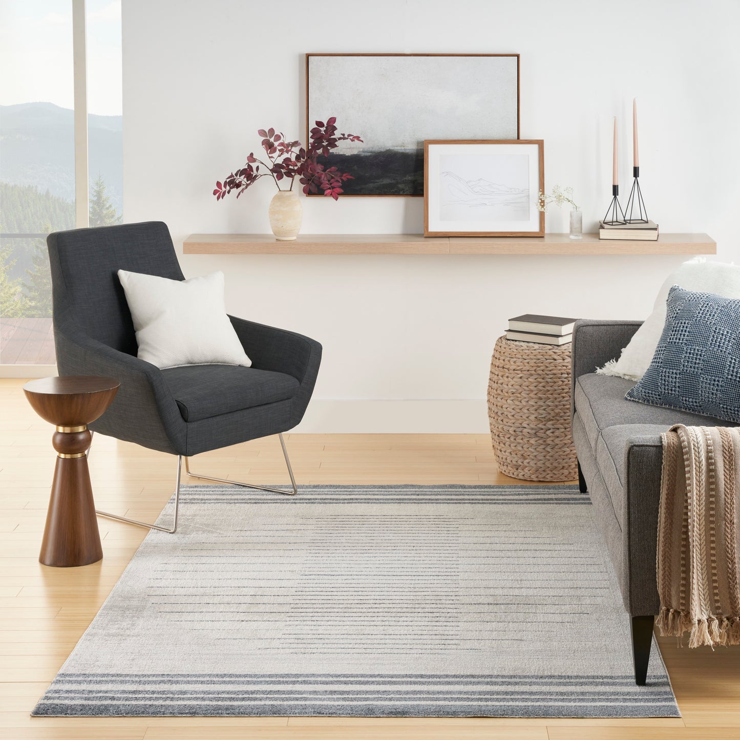 Dark gray rug 5' x 7' on a living room floor, with a gray sofa and blue cushions, a woven pouf, and a wooden shelf with framed art.