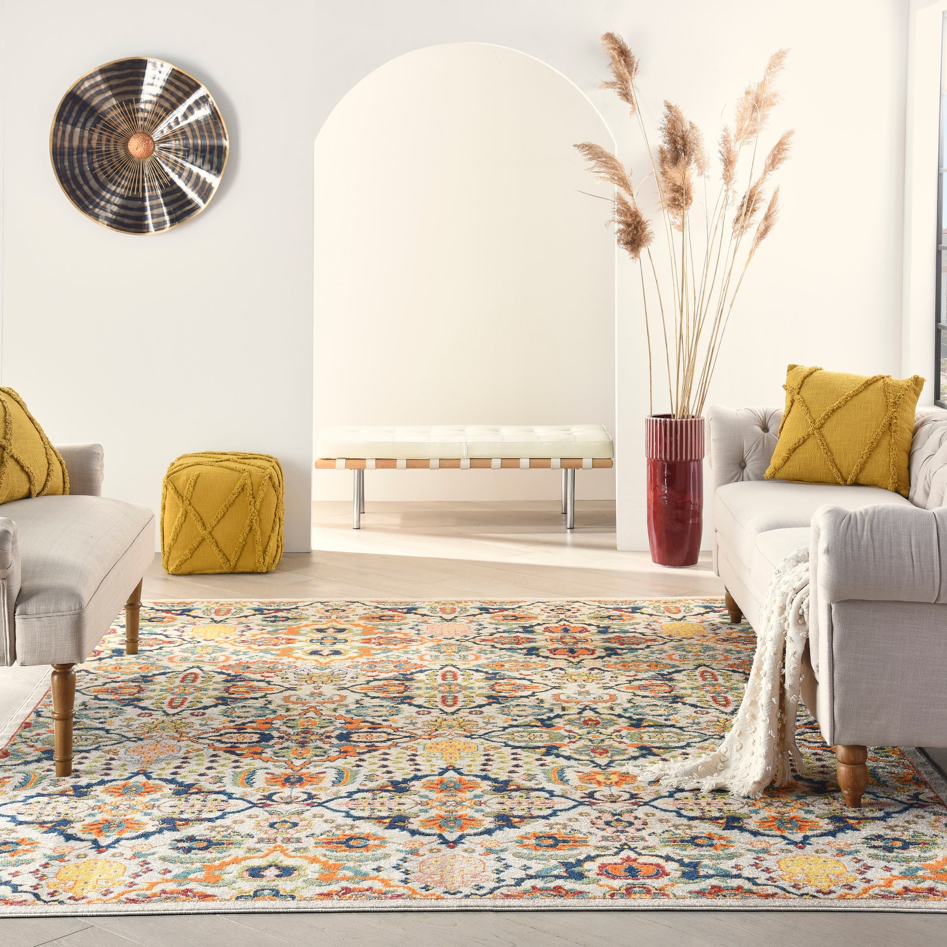 Cozy living room with blue-orange rug, beige sofas, mustard cushions, a white arched bench, and pampas grass in a red vase.