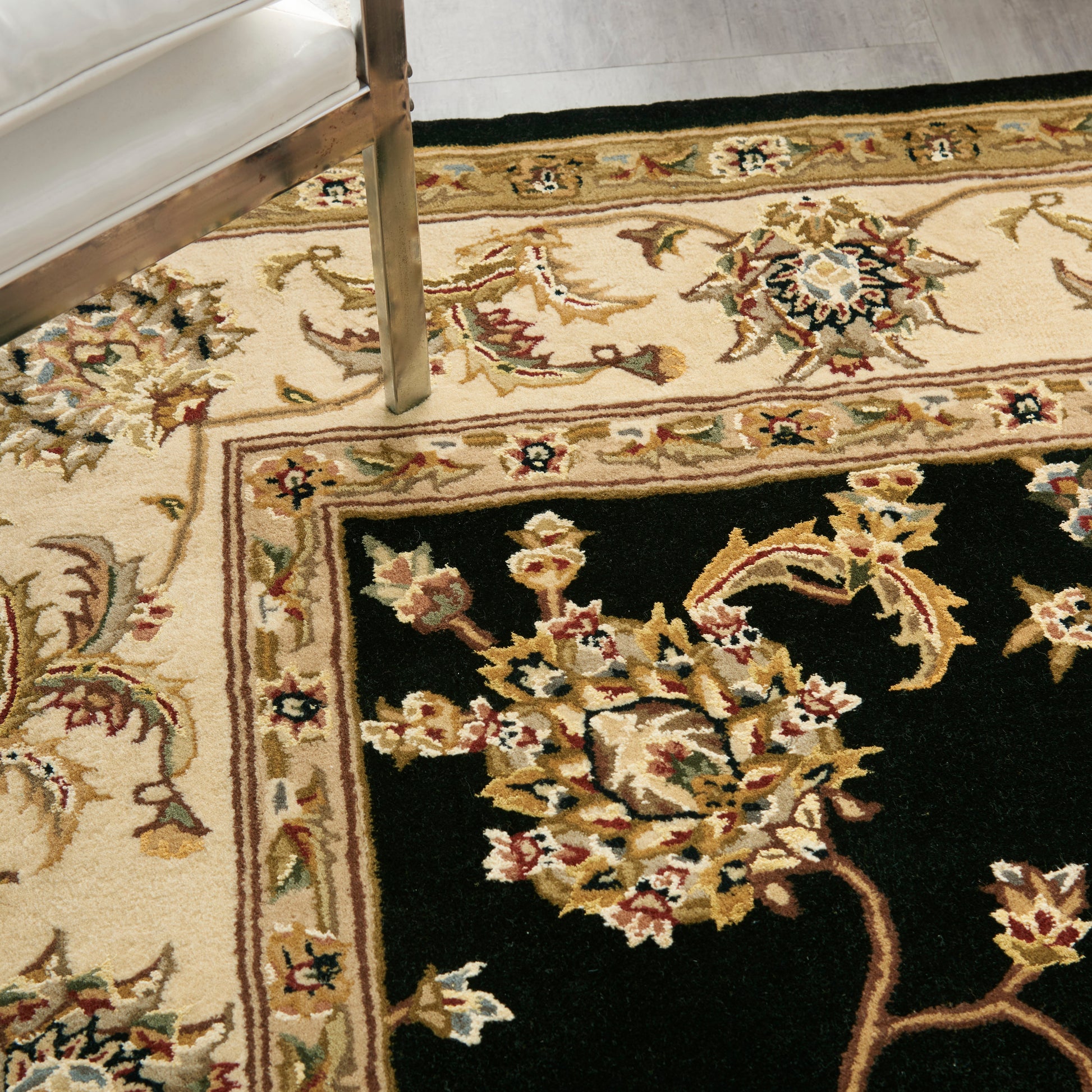 Traditional floral area rug: cream border with vines and blossoms, black panel with gold-pink florals; chair leg visible.