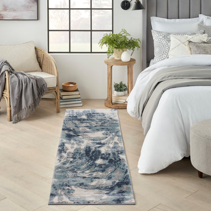 Blue abstract runner rug on light wood floor beside a bed with white bedding; rattan chair and wooden side table with plants, airy modern bedroom.