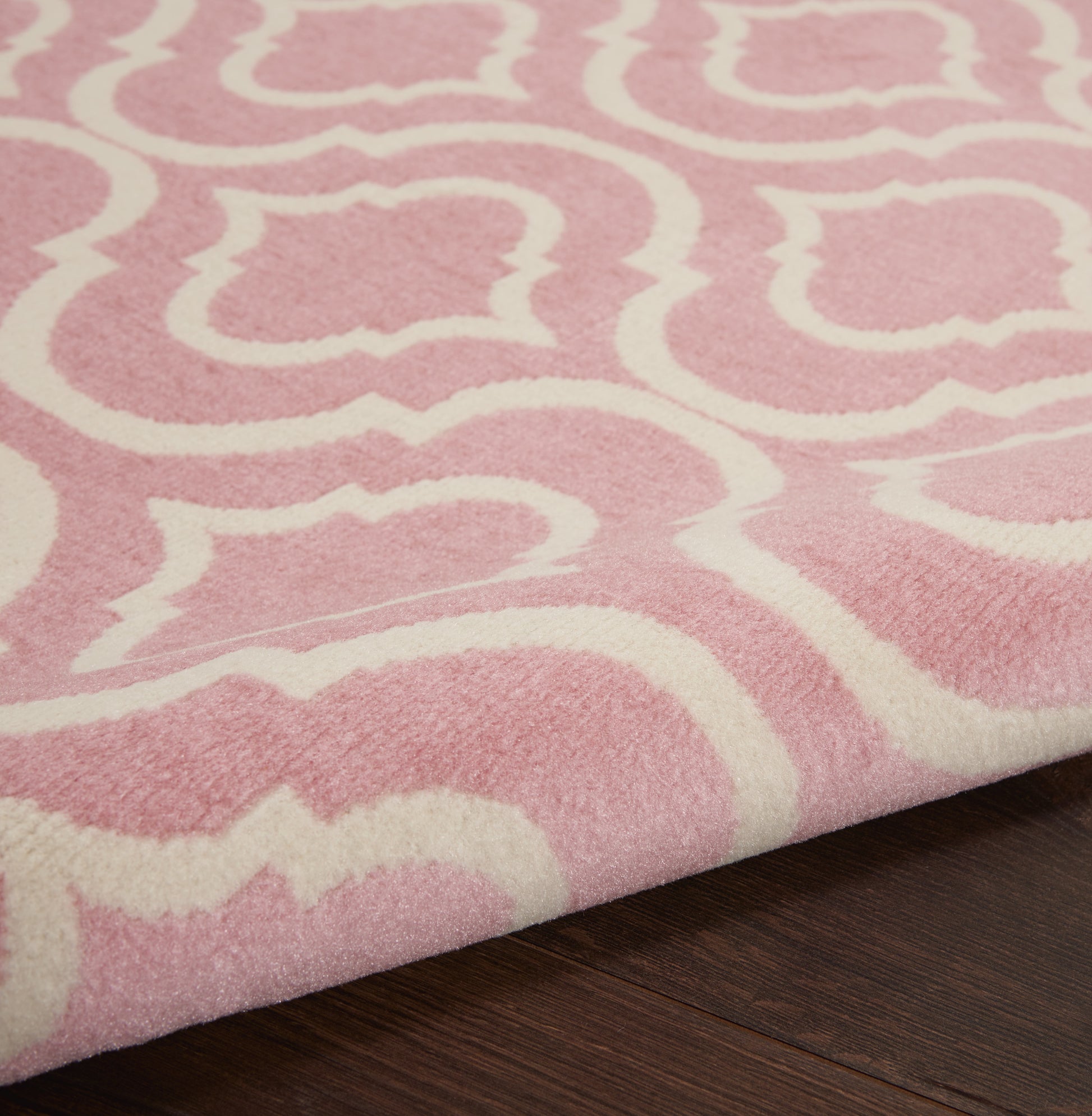 Pink plush rug with a white arabesque pattern on dark wood floor; soft pile with a folded corner.