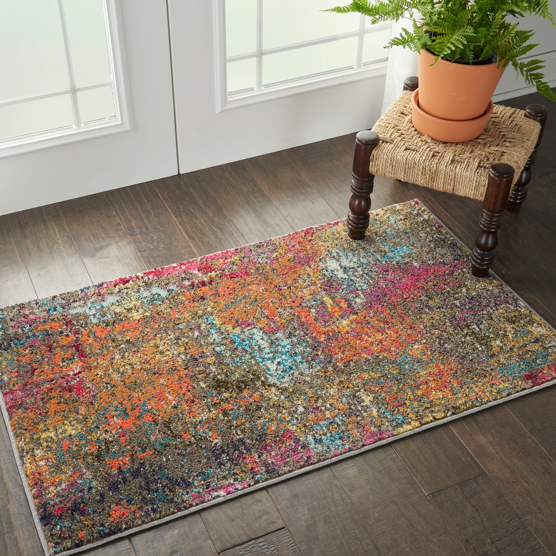 Rectangular multicolor abstract rug on dark wood floor beside a woven-stool with a potted fern.