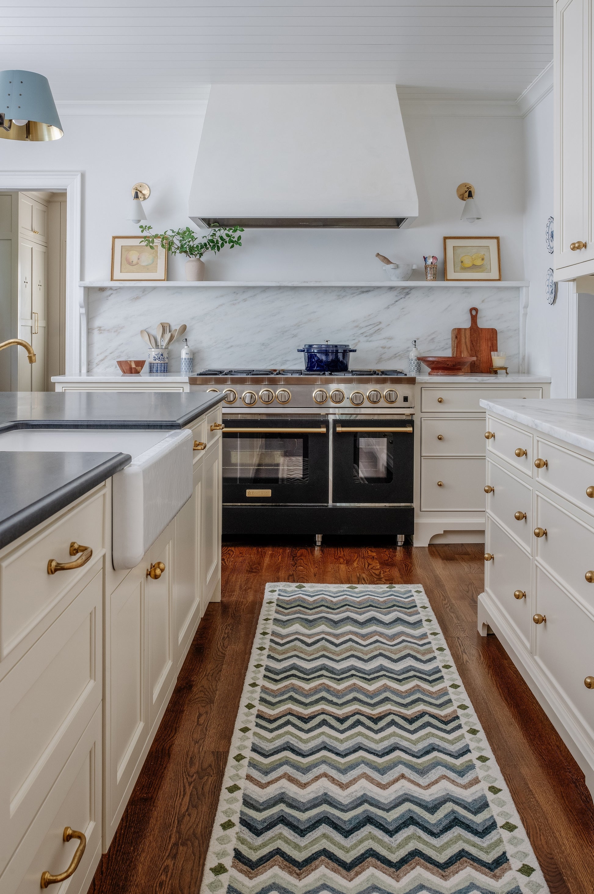White shaker kitchen with marble backsplash, black double-oven range, large white hood, brass hardware, blue pot, zigzag rug.