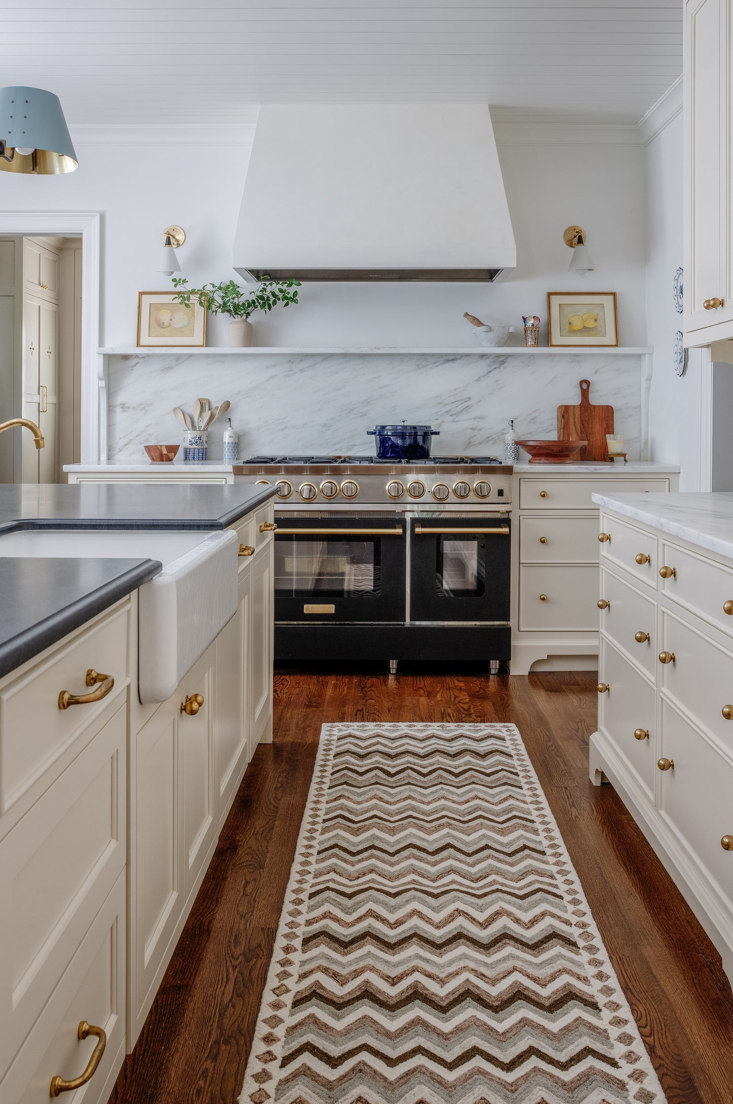 Bright white kitchen with marble backsplash, brass hardware, double-oven range, white hood, blue pendant light, chevron rug.