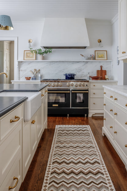 Bright white kitchen with marble backsplash, brass hardware, double-oven range, white hood, blue pendant light, chevron rug.