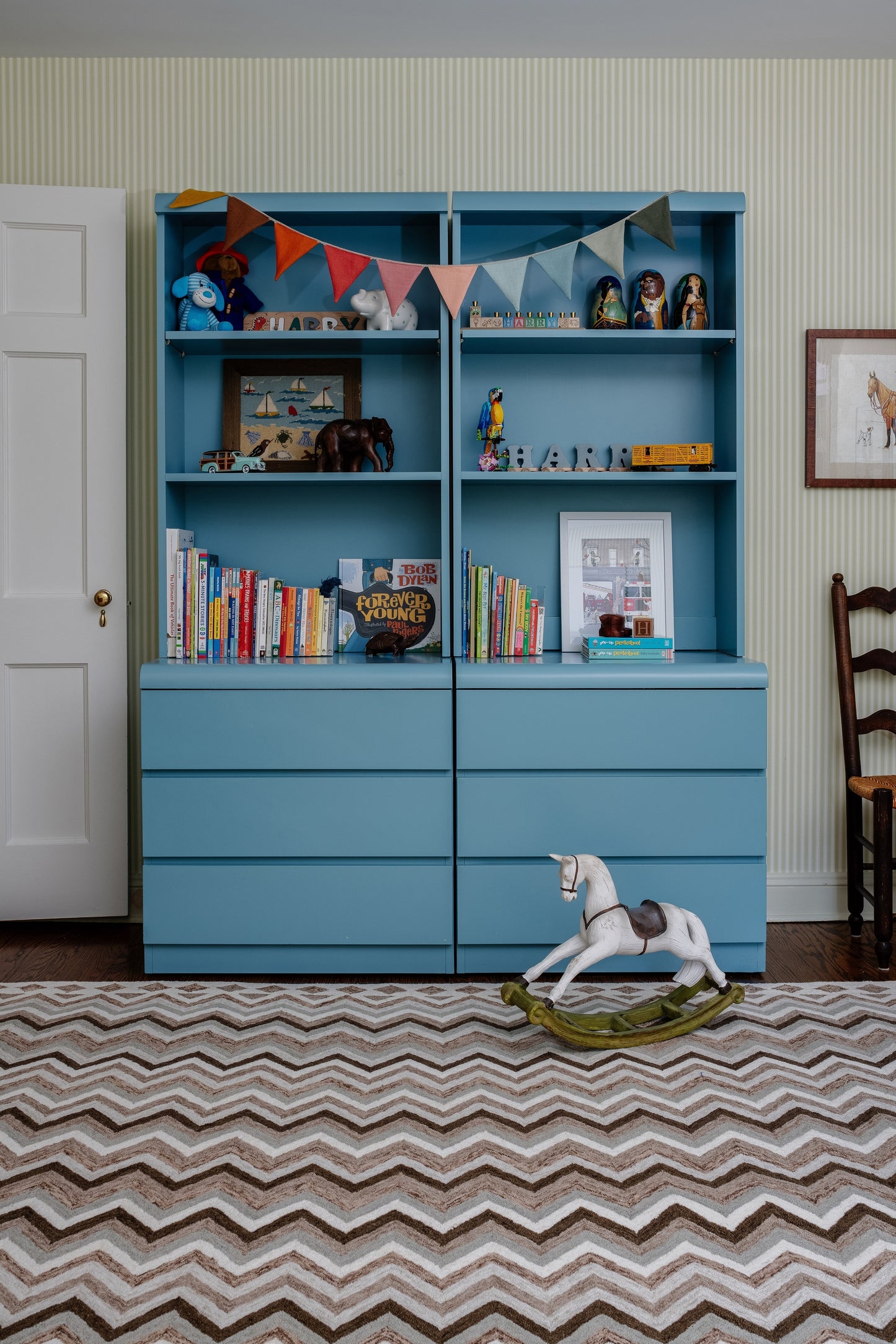 Blue wooden storage unit with open upper shelves and lower drawers, bunting, books, and a rocking horse on a chevron rug.
