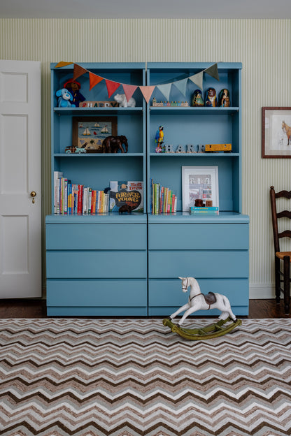 Blue-painted wooden storage unit with top shelves and bottom drawers, filled with books, decor, bunting, and a rocking horse.