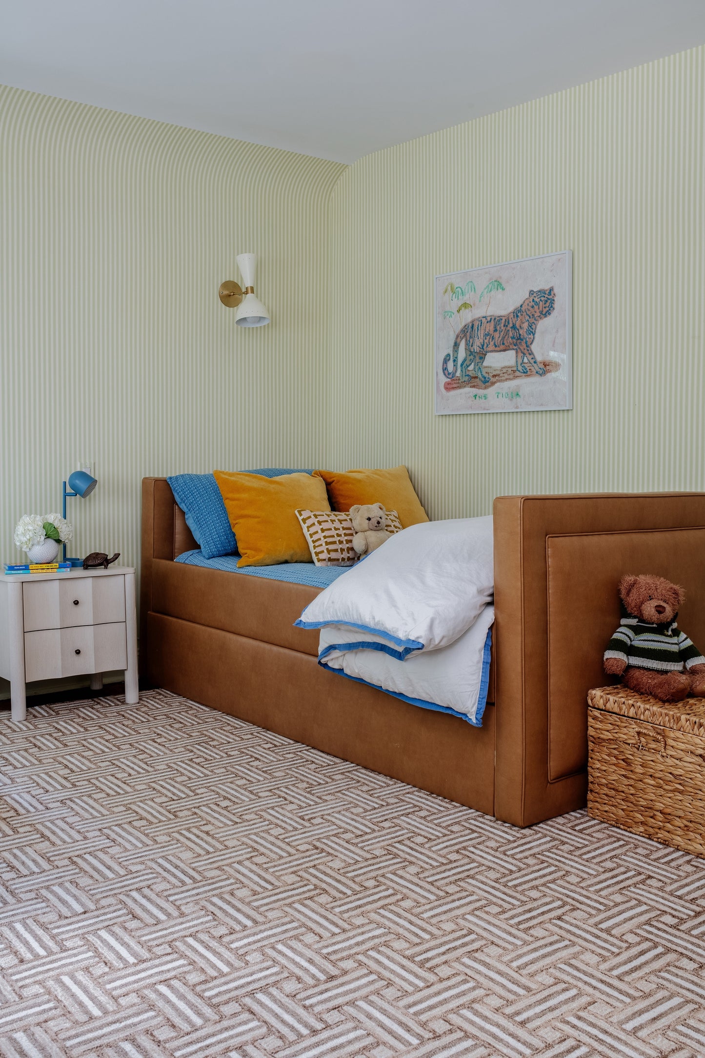 Brown upholstered kids' bed with blue and orange pillows, white bedding, tiger wall art, basket with teddy, striped walls
