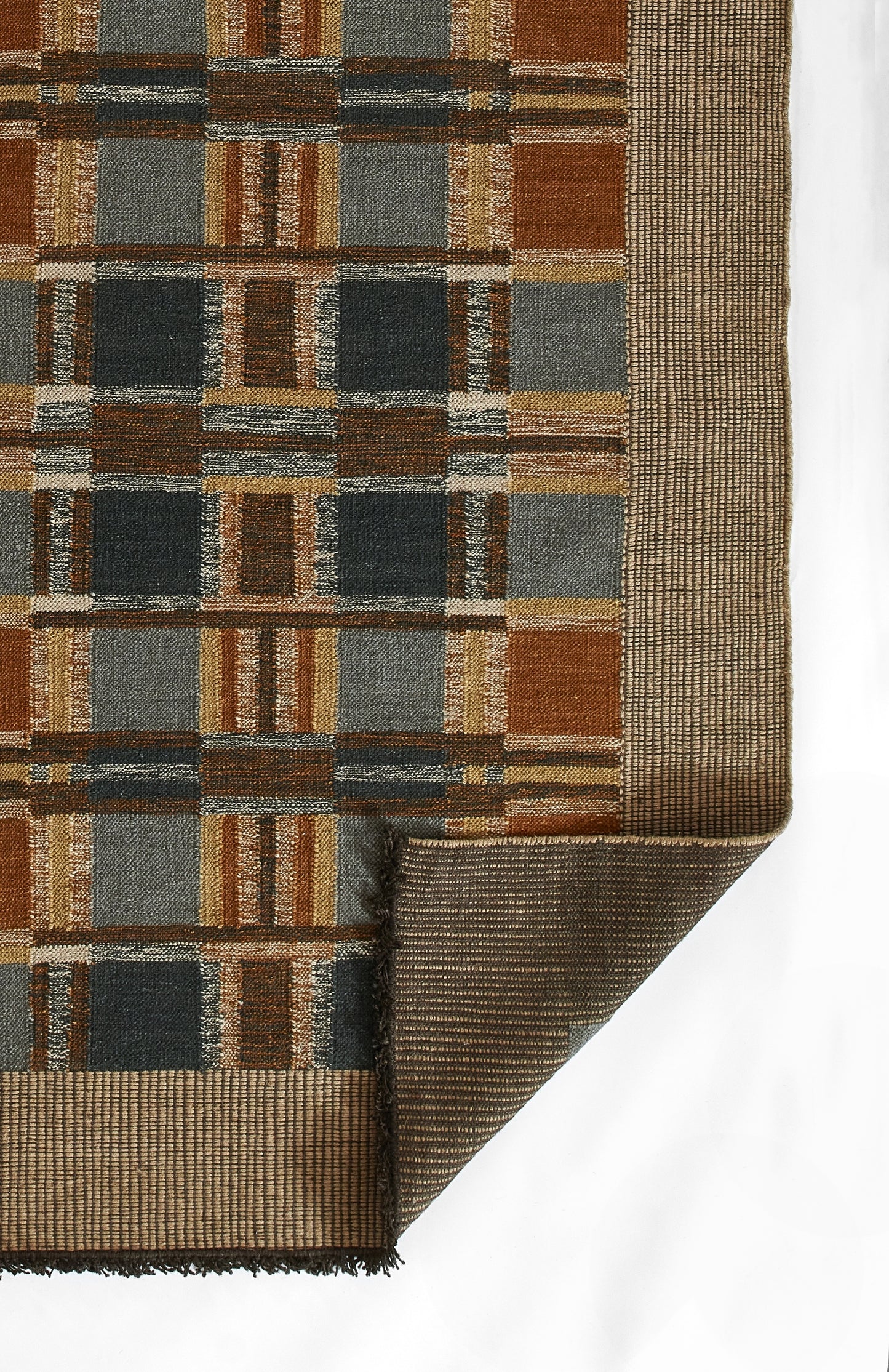 Geometric woven rug with brown, tan, rust, blue and gray blocks; folded corner reveals underside.