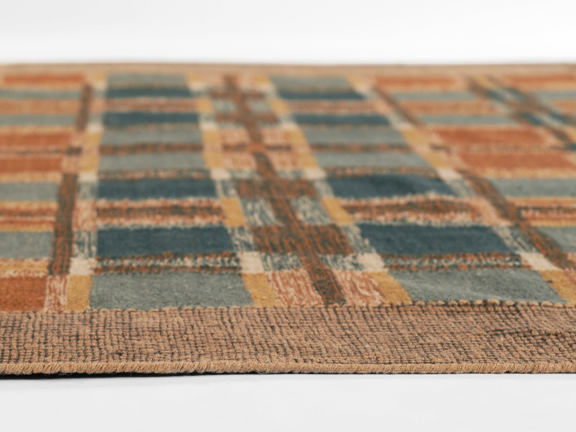 Close-up of a woven plaid rug in teal, burnt orange, and tan with a textured beige edge.