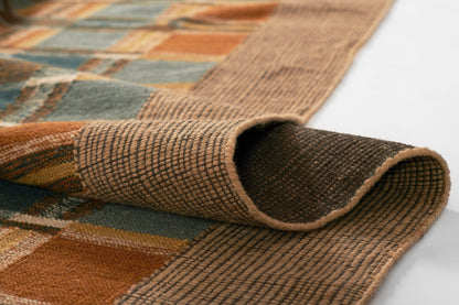 Woven plaid rug in blue, orange, and brown tones; textured flat-weave with tan edging and folded corner.